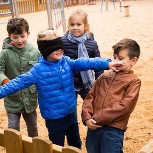 Geleneksel çocuk oyunlarından körebe oynayan çocuklar ve gözleri kapalı arkadaşlarını arayan mavi montlu sarışın erkek çocuk