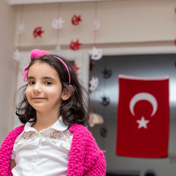 23 Nisan Ulusal Egemenlik ve Çocuk Bayramı'nda cama asılan Türk Bayrağı önünde gülümseyen, pembe tac, yelek ve beyaz gömlekli kız çocuğu