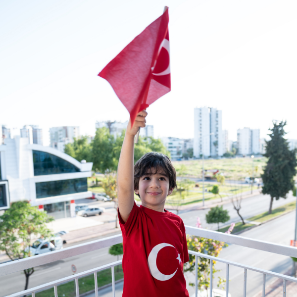 Şehir manzaralı bir balkonda, 23 Nisan Ulusal Egemenlik ve Çocuk Bayramı'nda elinde Türk Bayrağı tutan, gülümseyen Türk Bayrağı tişörtlü erkek çocuk