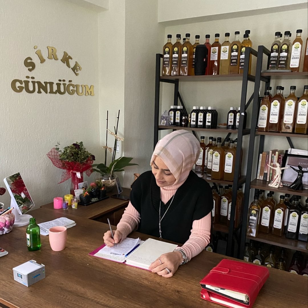 Feyza Hanım, Sirke Günlüğüm atölyesinde reçete defterine not alırken, arkasında raflarca doğal fermente sirke şişeleri