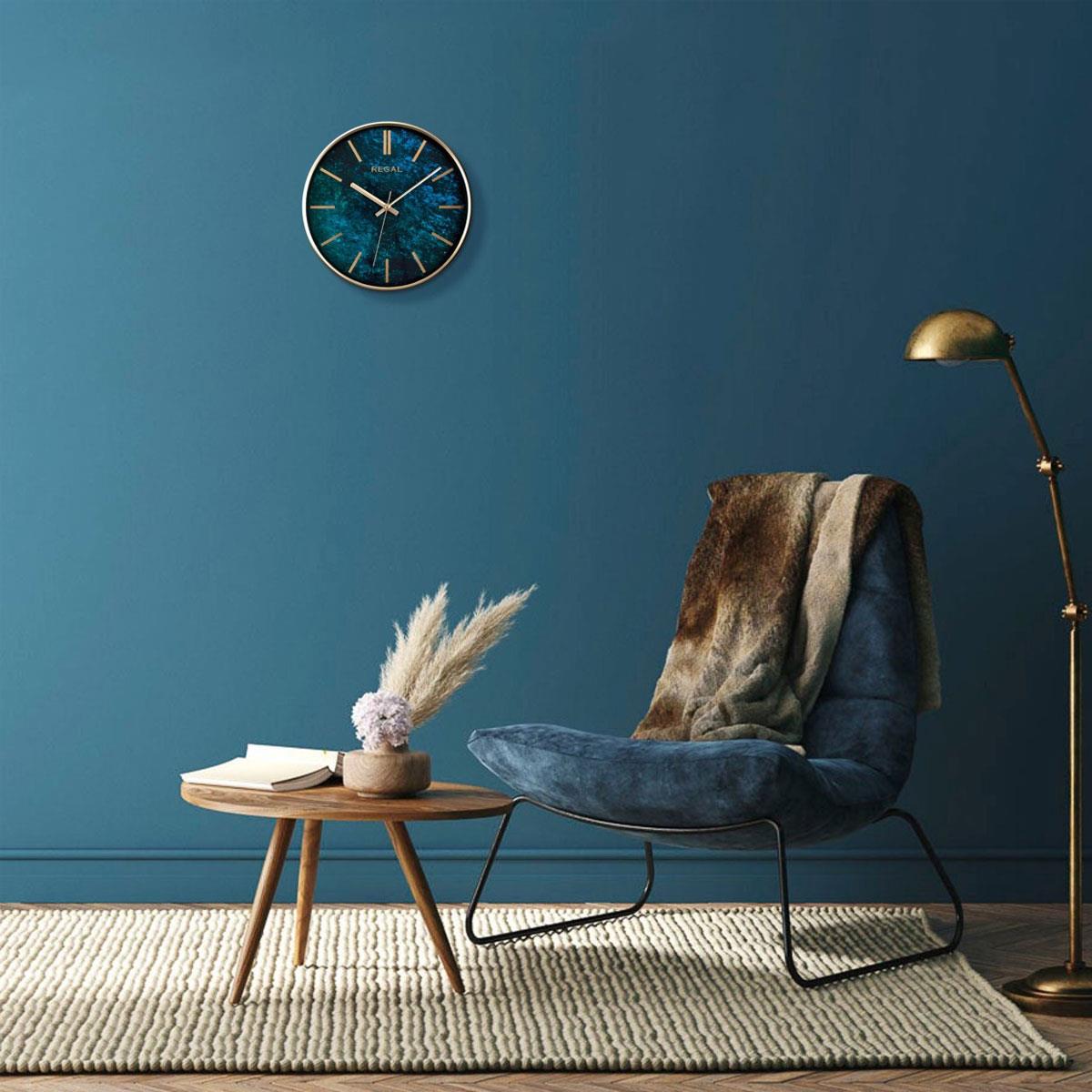 Modern bir yaşam alanında, mavi bir koltuk ve altın rengi lambanın yanında duvarda asılı teal desenli Regal duvar saati.